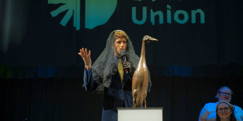 Greg James is wearing a black veil and speaking into a microphone, in front of the bronze statue of Long Boi. In the background is a cloak reading 'Students Union'